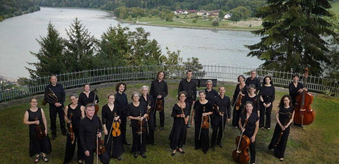 A group of musicians is standing outdoors, surrounded by trees and a river in the background. They are wearing black clothing and holding their instruments. | © Reinhard Winkler