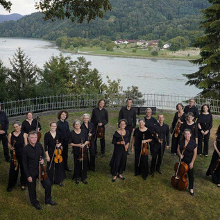 A group of musicians is standing outdoors, surrounded by trees and a river in the background. They are wearing black clothing and holding their instruments. | © Reinhard Winkler