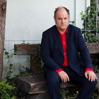 Egy férfi egy fapadon ül egy fa alatt. Sötét öltönyt és piros inget visel, miközben gondolkodva néz a kamerába. | © Stephan Minx