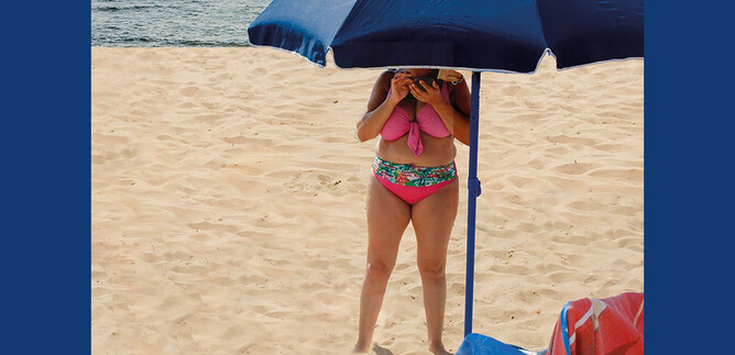 A person is standing on the beach under a sun umbrella. The sand is light and the sea is visible in the background. | © Valentina Schuster