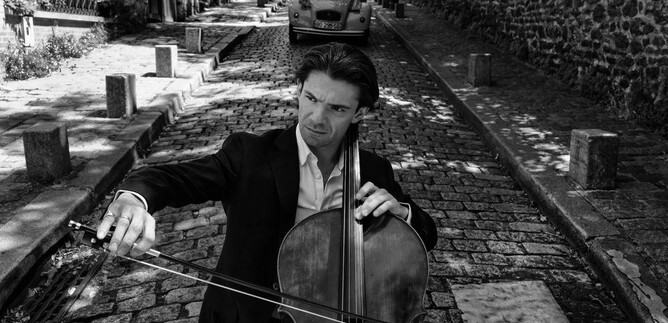 Un violoncelliste joue sur une rue pavée dans une ville pittoresque. En arrière-plan, on peut voir une vieille voiture et des bâtiments caractéristique. | © Anoush Abrar