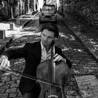 A cellist is playing on a cobblestone street in a picturesque town. In the background, an older car and characteristic buildings can be seen. | © Anoush Abrar