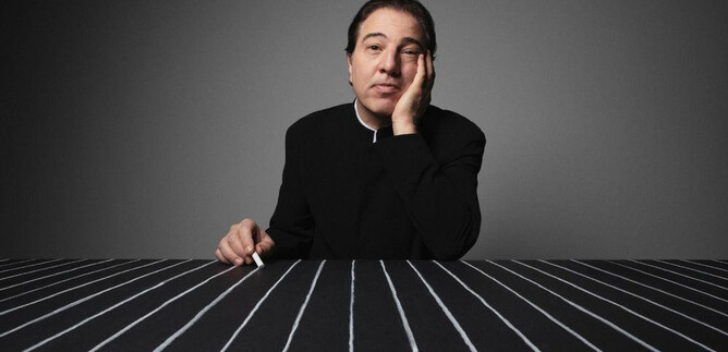 Ein nachdenklicher Mann sitzt an einem Tisch mit schwarz-weißen Streifen. Er hat eine Hand an sein Gesicht gelegt und sieht ernst aus. | © Fethi Karaduman