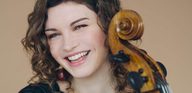 Une femme souriante avec des cheveux bouclés tient un violoncelle. Le fond est dans des tons terre doux. | © Simon Pauly