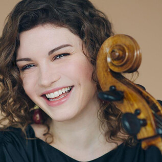 Usměvavá žena s kudrnatými vlasy drží violoncello. Pozadí je v jemných zemních tónech. | © Simon Pauly