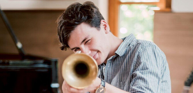 Un jeune homme joue avec enthousiasme d'une trompette. En arrière-plan, on peut voir un piano et une atmosphère chaleureuse et amicale dans la pièce. | © Kunsthilfe Salzburg