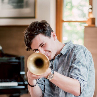 A young man is enthusiastically playing a trumpet. In the background, a piano and a warm, friendly room atmosphere can be seen. | © Kunsthilfe Salzburg