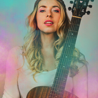 Egy fiatal nő, aki göndör szőke hajú, egy akusztikus gitárt tart a kezében. Magabiztos mosolya van, és egy egyszerű felsőt visel. | © Christian Verch