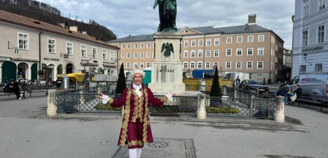 A person in historical clothing stands in front of a monument in a city square. Buildings and a graceful sky can be seen in the background. | © Sabine Rath