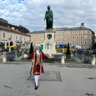 A person in historical clothing stands in front of a monument in a city square. Buildings and a graceful sky can be seen in the background. | © Sabine Rath