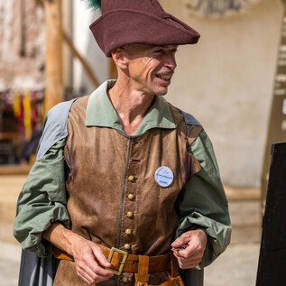 Un homme en costume historique se tient souriant dans un environnement médiéval. Il porte un chapeau brun avec une plume et a une sacoche en cuir sur le côté.