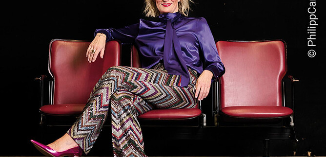 A stylishly dressed woman sits relaxed on a red theater chair. She is wearing a shimmering top and patterned pants. | © PhilippCarlRiedl