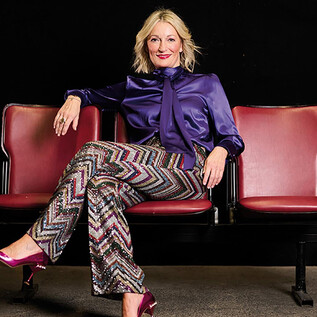 A stylishly dressed woman sits relaxed on a red theater chair. She is wearing a shimmering top and patterned pants. | © PhilippCarlRiedl