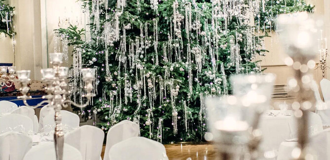 An elegant banquet hall with a decorated Christmas tree in the background. Tables are adorned with white tablecloths and candle holders. | © St. Peter Stiftskulinarium