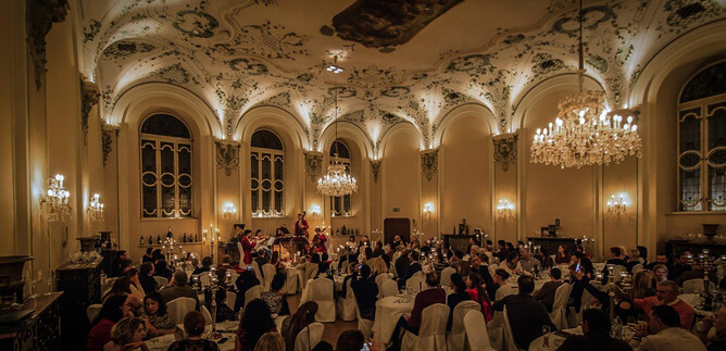 An elegant event space with many guests sitting at festively set tables. The room is atmospherically lit and adorned with magnificent chandeliers. | © Michael Groessinger