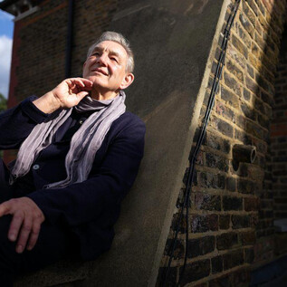 An older man is sitting relaxed on a wall and smiling. He is wearing a scarf and has a thoughtful pose. | © Gerard Collet