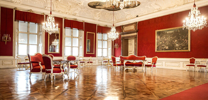 Egy elegáns szoba piros falakkal és luxus bútordarabokkal. Kristálycsillárok lógnak a mennyezetről, és megvilágítják a fényes padlót. | © DomQuartier