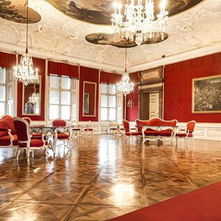 An elegant room with red walls and luxurious furniture. Crystal chandeliers hang from the ceiling and illuminate the glossy floor. | © DomQuartier
