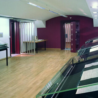 A modern exhibition room with wooden floors and bright walls. Glass display pieces are placed along the walls. | © ISM