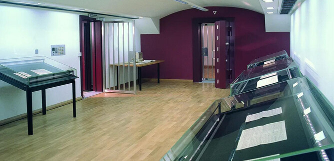 A modern exhibition space with wooden flooring and red walls. Glass cases showcase various documents and exhibits. | © ISM