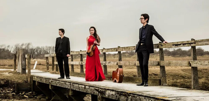 Three musicians are standing on a wooden walkway, surrounded by an autumn landscape. The woman is wearing a red dress and holding a musical instrument in her hand. | © Foppe Schut