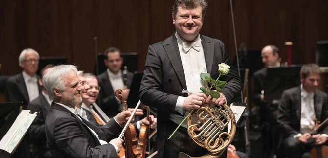 A musician is smiling on stage with a horn and a rose. In the background, other orchestra musicians can be seen. | © Wolfgang Lienbacher