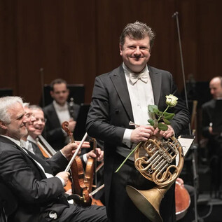 A musician is smiling on stage with a horn and a rose. In the background, other orchestra musicians can be seen. | © Wolfgang Lienbacher