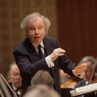 A conductor leads an orchestra with passionate gestures. In the background, musicians with their instruments can be seen. | © Priska Ketterer