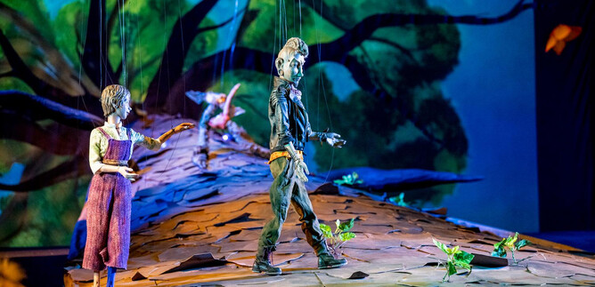A stage with two puppets standing on a large tree trunk. In the background, there is a vibrant forest scene with colorful hues. | © Bernhard Mueller