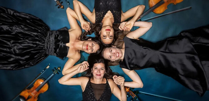 Quattro donne in eleganti abiti neri sono sdraiate a terra e si tengono per mano. Intorno a loro sono posizionati dei violini. | © Ernest Stierschneide