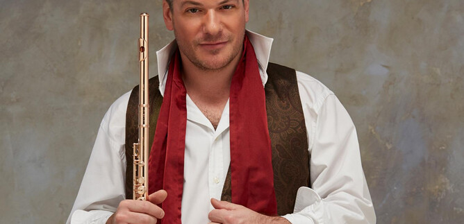 Ein Mann hält eine Flöte in der Hand und trägt ein elegantes Outfit mit einem roten Schal. Der Hintergrund ist neutral und leicht strukturiert. | © Fabien Monthubert