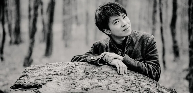 Ein junger Mann lehnt sich an einen Baumstamm im Wald. Die Szene ist in Schwarz-Weiß gehalten und vermittelt eine ruhige Atmosphäre. | © Marco Borggreve