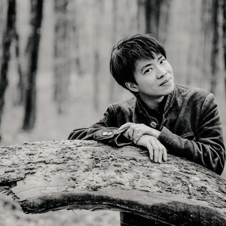 A young man leans against a tree trunk in the forest. The scene is in black and white and conveys a calm atmosphere. | © Marco Borggreve