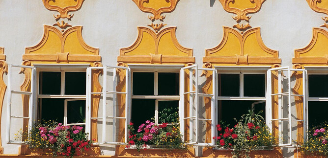 Kolorowa elewacja domu z żółtym tynkiem i otwartymi oknami. Parapety są ozdobione kwitnącymi roślinami. | © TSG