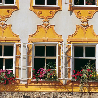 A colorful house facade with yellow plaster and open windows. The window sills are adorned with blooming plants. | © TSG
