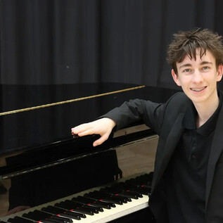 A young man is sitting at a piano and smiling. He is wearing a black outfit and is standing in front of a black, shiny piano. | © Moriz Purgstaller