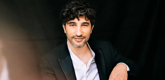 Egy férfi fekete öltönyben ül egy sötét háttér előtt. Barátságosan mosolyog és göndör haja van. | © Christoph Köstlin