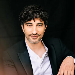 A man in a black suit is sitting in front of a dark background. He is smiling warmly and has curly hair. | © Christoph Köstlin
