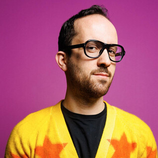 A man with glasses and a colorful sweater is standing in front of a pink background. He is looking thoughtfully at the camera. | © Peter Rigaud