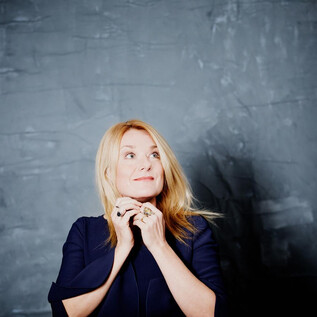 Eine Frau mit blonden Haaren schaut nachdenklich nach oben. Sie trägt ein dunkelblaues Oberteil vor einer grauen Wand. | © Julia Wesely