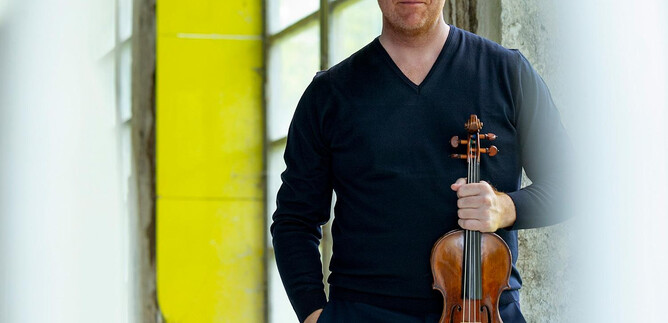 A man stands in a room holding a violin in his hand. The background is bright with large windows. | © Inge Prader