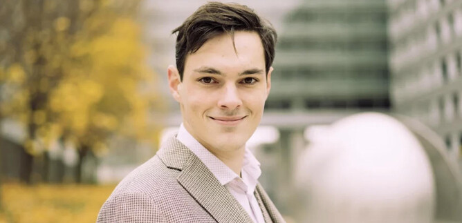 A young man in an elegant suit smiles warmly at the camera. In the background, modern buildings and autumn trees can be seen. | © Lukas Beck