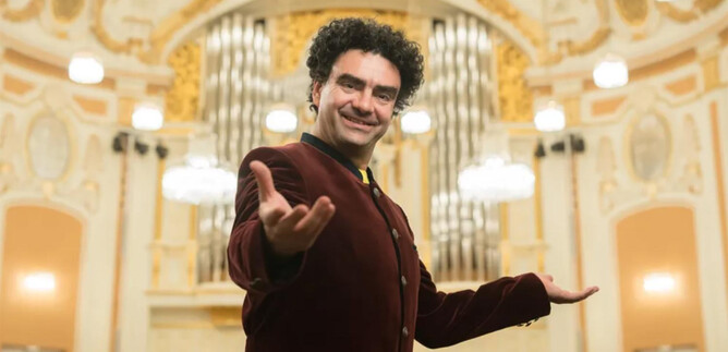 A man in an elegant red jacket stands in front of an impressive organ in a festive hall. He is smiling and seems to be welcoming the audience. | © Wolfgang Lienbacher