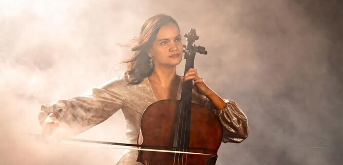 Eine junge Frau spielt Cello in einem nebligen Umfeld. Sie trägt ein glitzerndes Outfit und schaut konzentriert. | © Michaela Weber