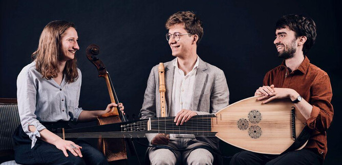 Tre musicisti sono seduti insieme e suonano diversi strumenti. Sorridono e godono dell'atmosfera. | © Johannes Berger