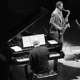 Une prise de vue en noir et blanc d'un pianiste jouant sur un piano, et d'un saxophoniste au premier plan. La scène est doucement éclairée, créant une atmosphère intime. | © Robert Lewis