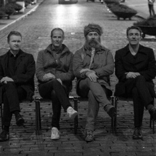 Four men are sitting on benches in an empty street at night. The scene is rendered in black and white and conveys a calm, nostalgic atmosphere. | © Knut Bry