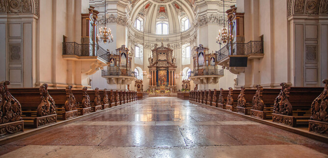 Une église impressionnante avec une haute voûte et des ornements artistiques. Les rangées de bancs en bois mènent à la magnifique zone de l'autel. | © Erzdiözese Salzburg