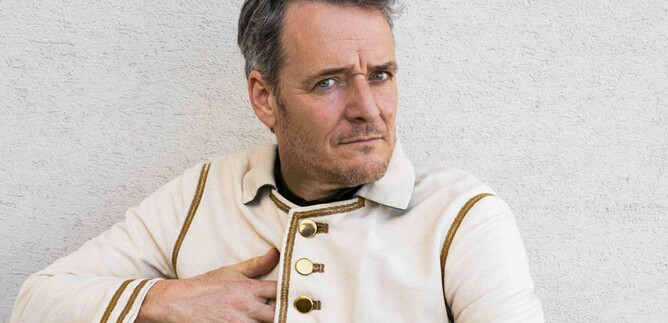 Egy férfi feltűnő fehér kabátban, aranygombokkal ül kényelmesen. Rövid szürke haja van, és közvetlenül a kamerába néz. | © Paul Vincent Schütz