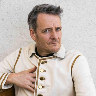A man in a striking white jacket with golden buttons sits comfortably. He has short gray hair and is looking directly at the camera. | © Paul Vincent Schütz